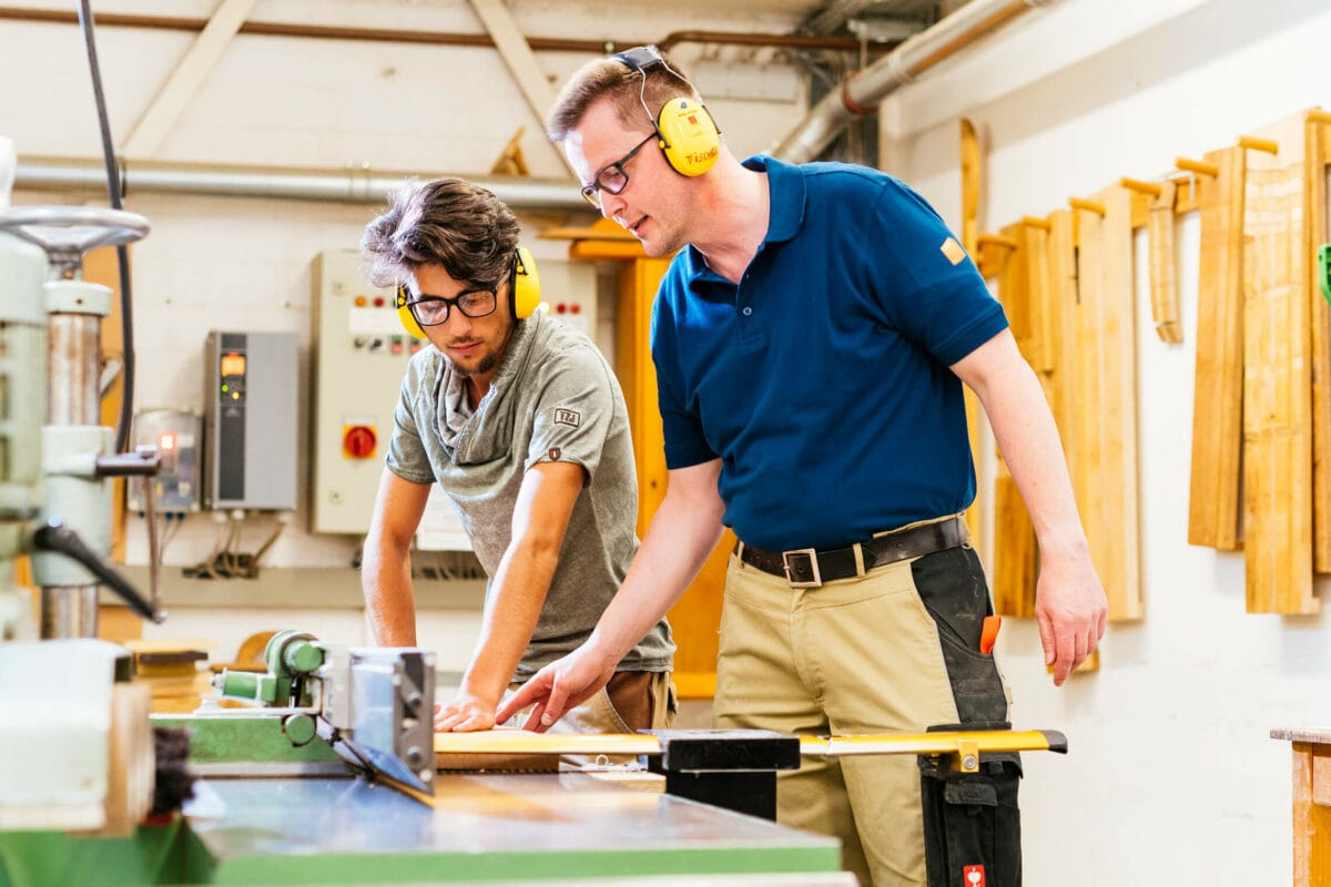 Ausbilder und Auszubildender mit Gehörschutz an einer Holzbearbeitungsmaschine in einer Tischlerei