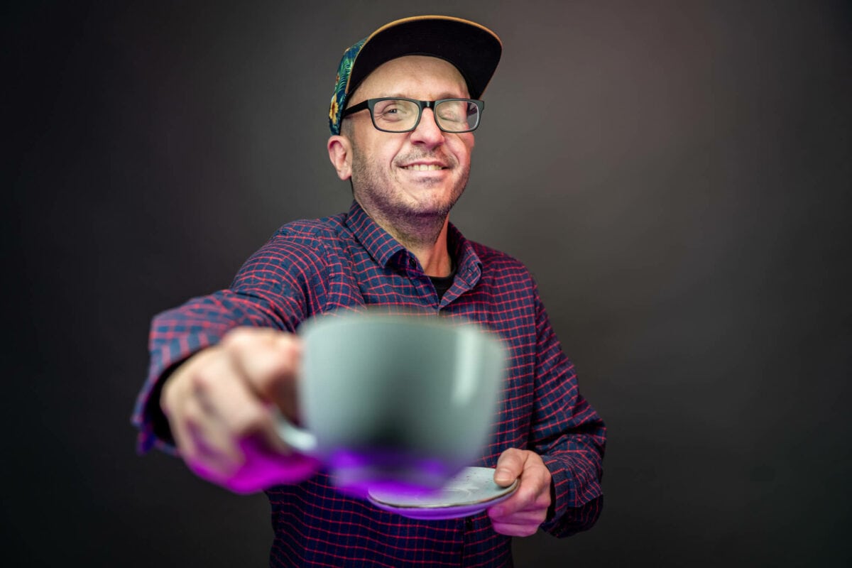 Mann mit Cap und Brille streckt lachend eine Kaffeetasse in die Kamera